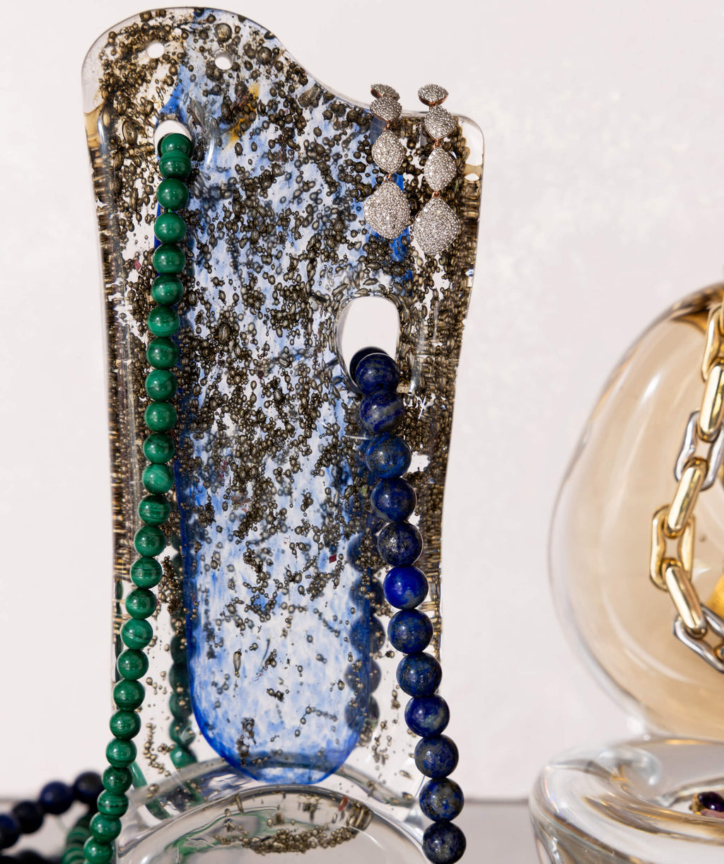 Close up of strings of lapis and malachite beads and diamond earrings  on a sculptural glass object
